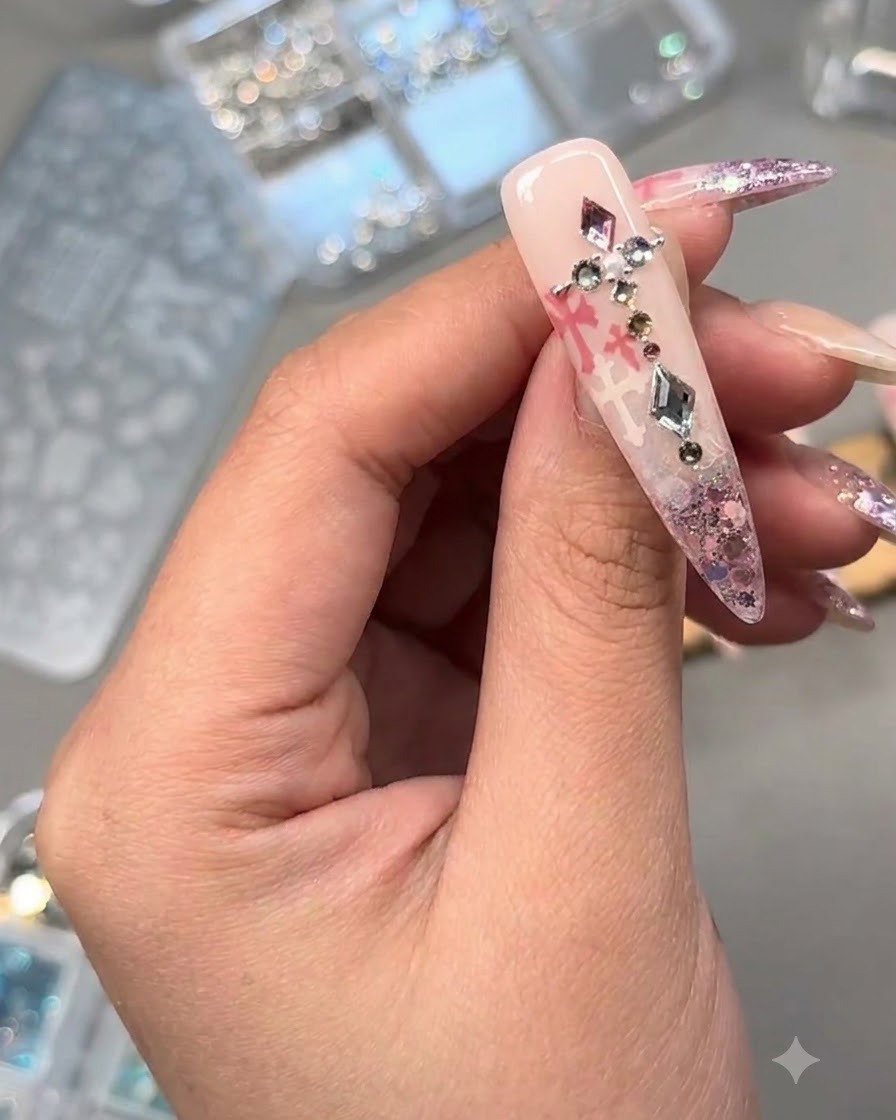 Gros plan dun ongle décoré orné de cristaux, dune perle et de charms métalliques sur fond de paillettes roses et dart estampé.
