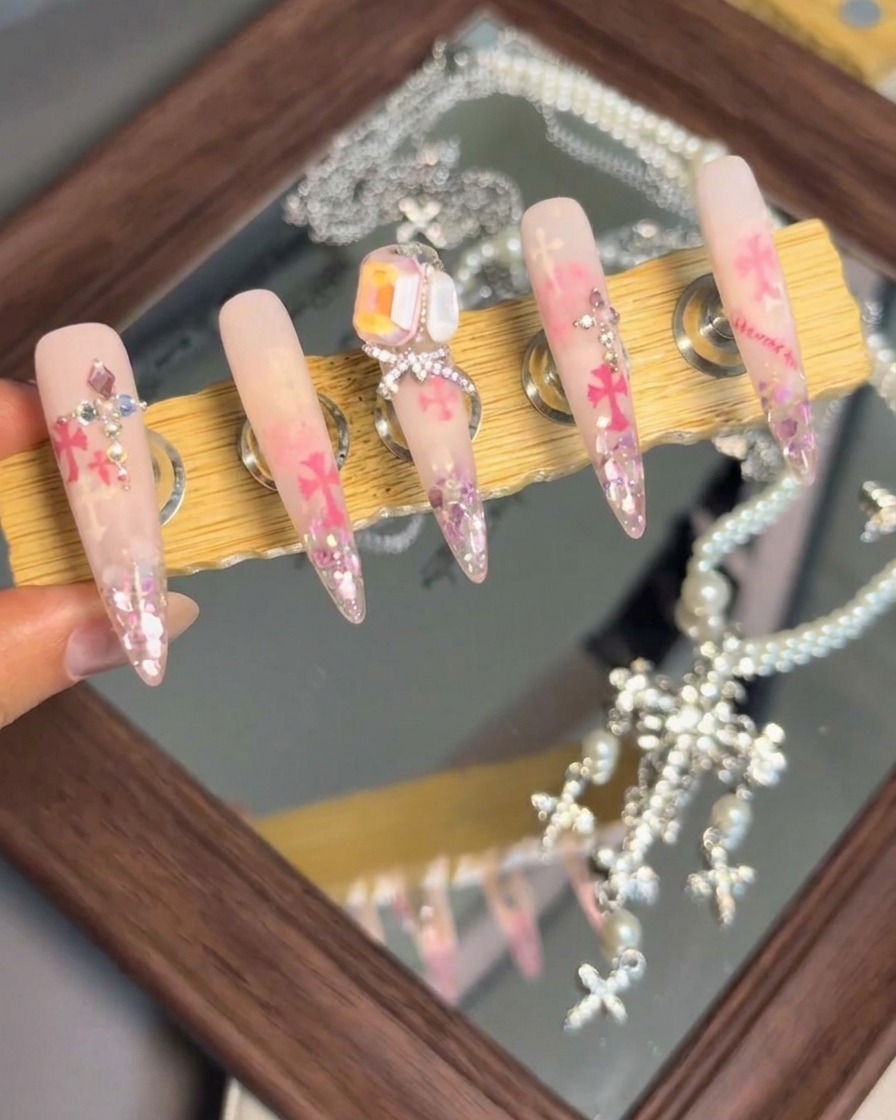 Un support présentant cinq faux ongles finis avec des agencements de cristaux élaborés, des nœuds et des accents Chrome Hearts.