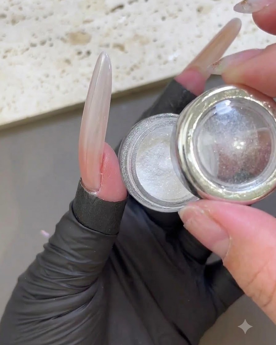 Une main gantée tenant un pot ouvert de poudre irisée blanche clair de lune à côté dun ongle rallongé brillant.