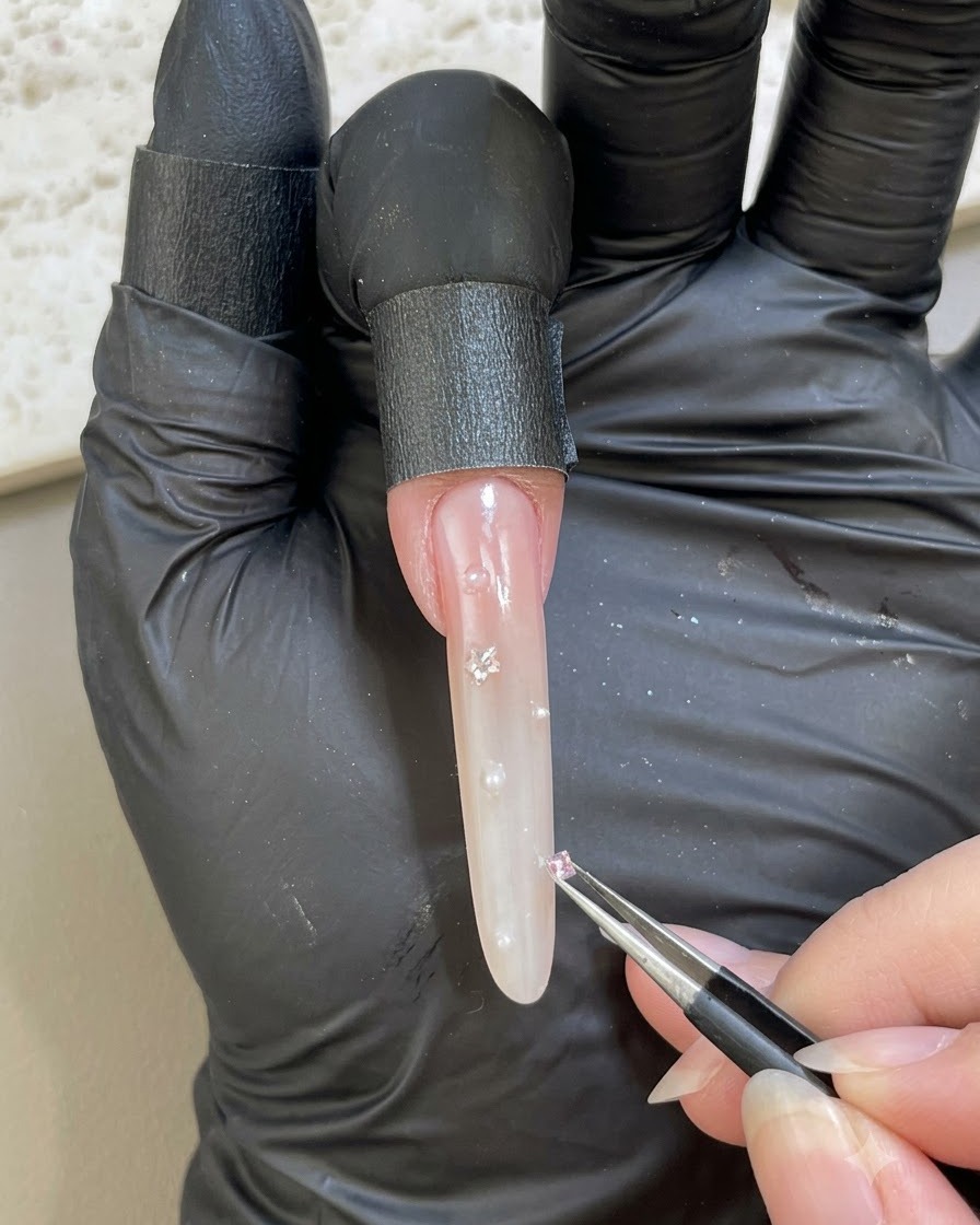 Une main gantée utilisant une pince pour placer un petit strass rose sur une surface dongle embellie.
