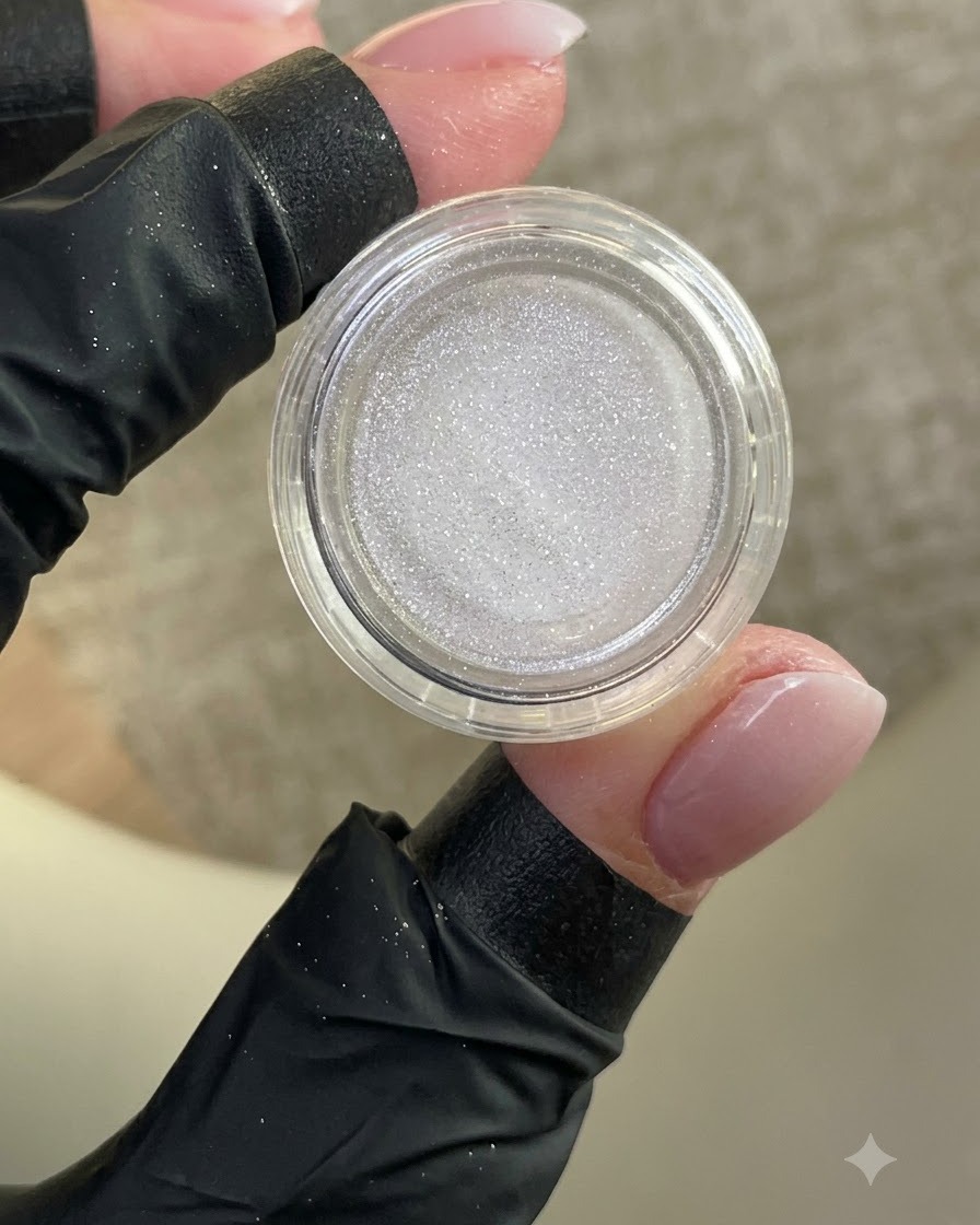 Une main gantée tenant un petit pot ouvert de poudre blanche chatoyante avec un ongle manucuré en vue.