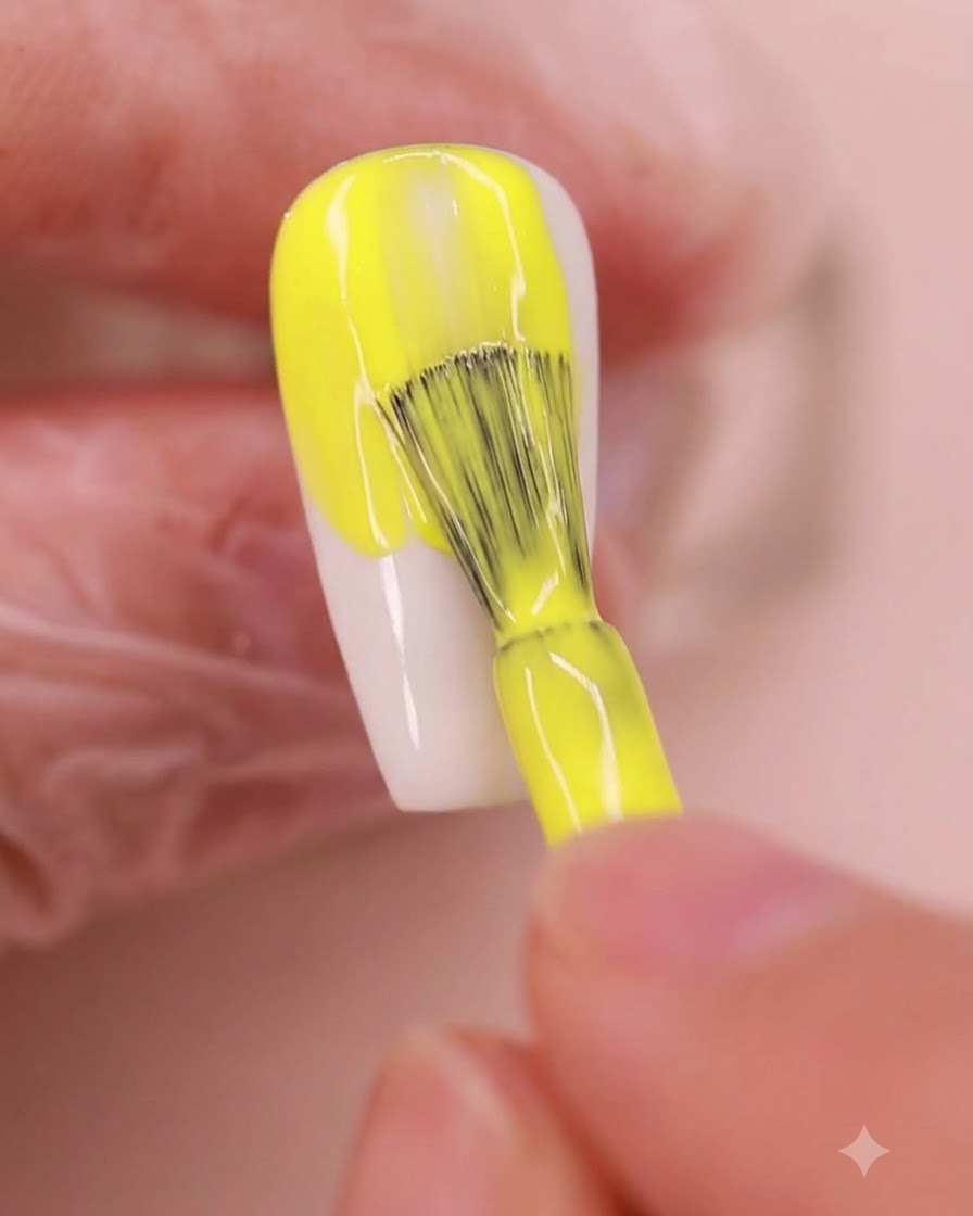 Un pinceau à ongle appliquant un gel polish jaune vif uniformément sur une plaque dongle en forme damande.