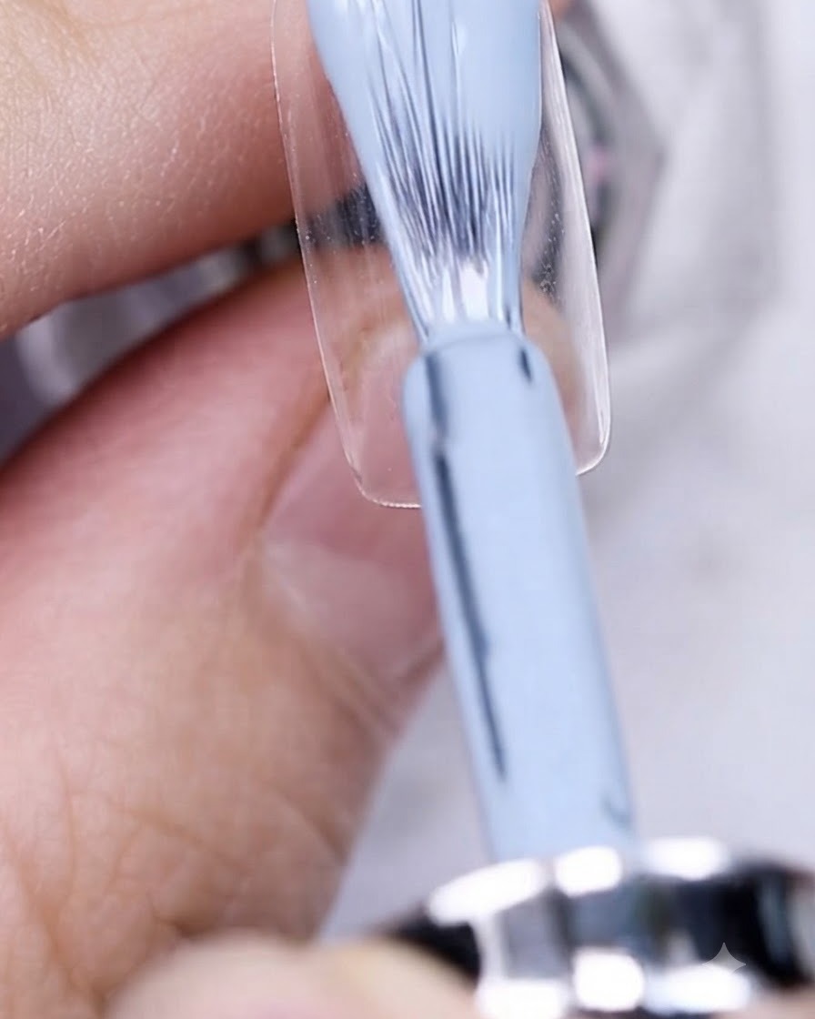 Un pinceau appliquant du vernis gel bleu grisé sur une plaque dongle préparée.
