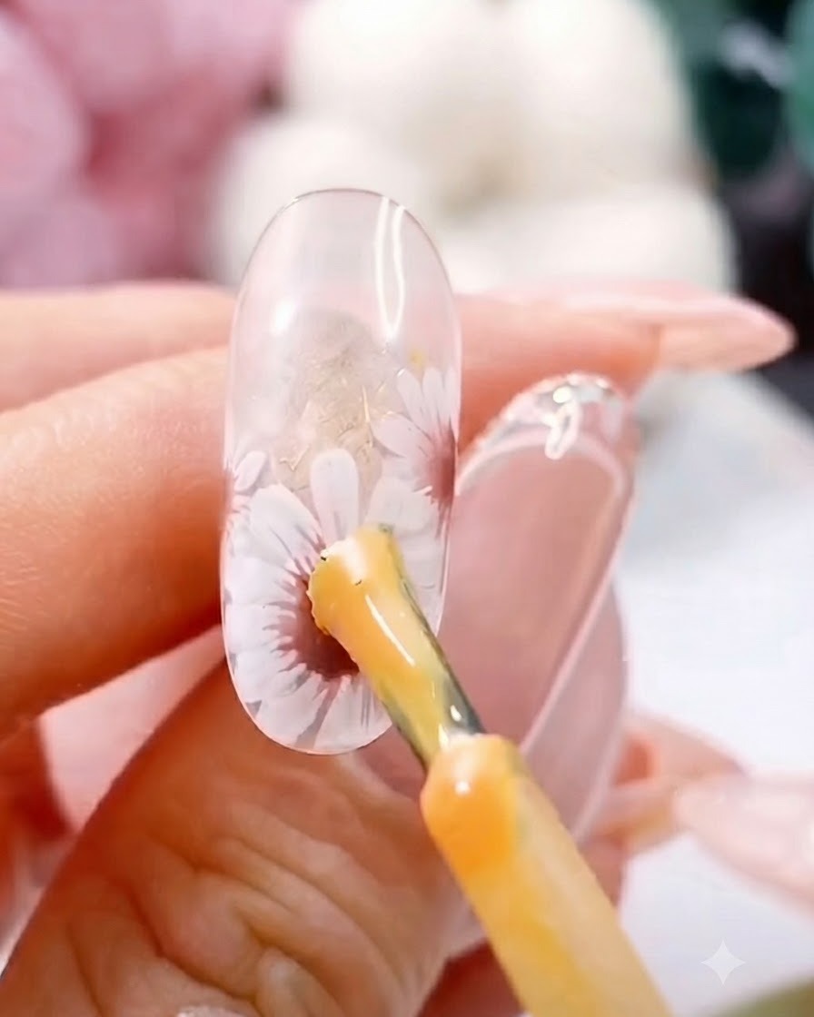 Un pinceau appliquant un point de gel jaune épais au centre dune marguerite blanche peinte sur un ongle transparent.