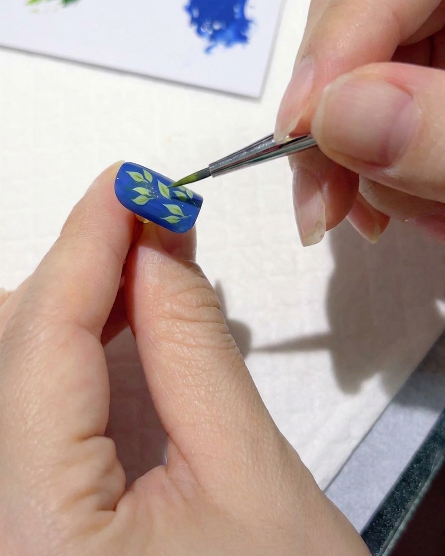 Un pinceau de nail art fin ajoutant des traits vert clair sur des feuilles sombres sur un ongle bleu.