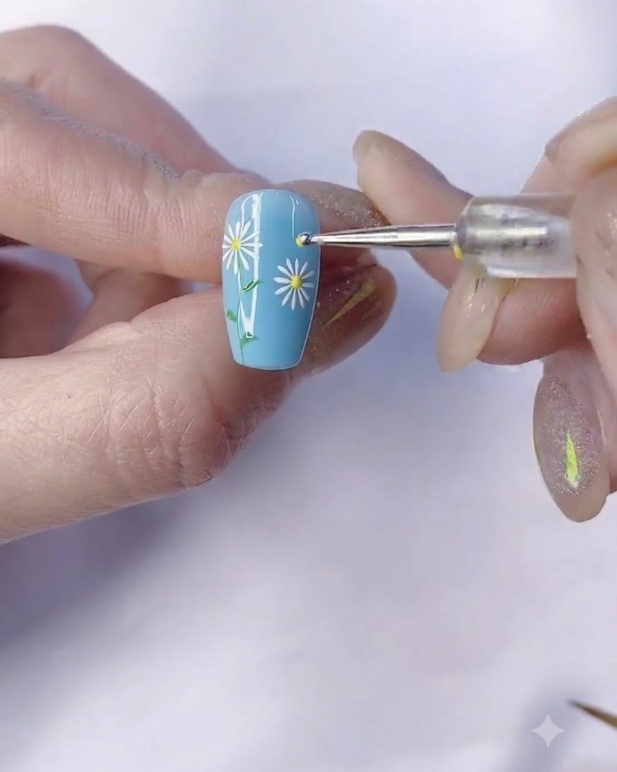 Un pinceau fin dessinant une tige verte partant de la marguerite sur un ongle bleu manucuré.