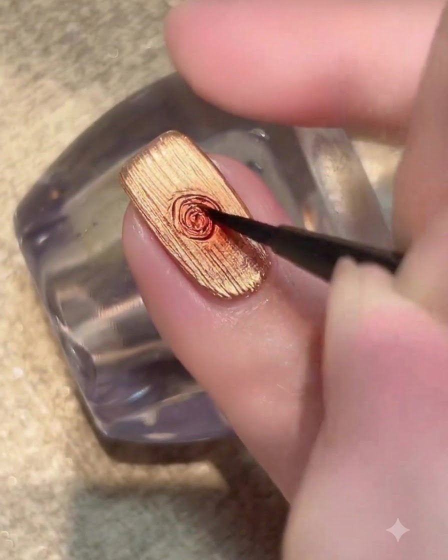 Un pinceau fin déposant une petite goutte de gel rouge foncé au milieu dun ongle doré.