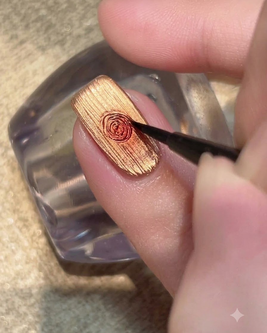 Un pinceau fin estompant une goutte de gel rouge en un motif de rose circulaire sur un ongle en or métallique.