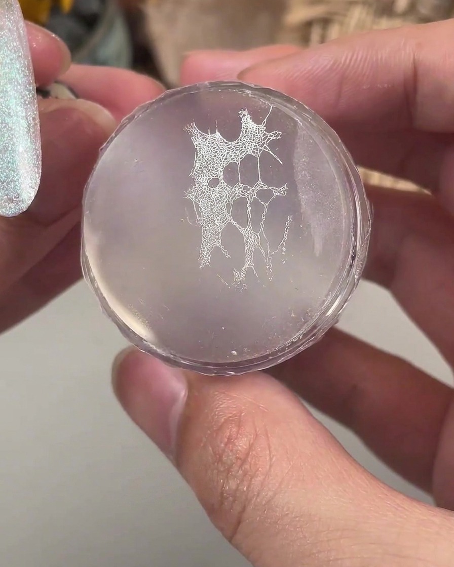 Un tampon en silicone transparent maintenant un motif de dentelle blanche fine prêt à être transféré sur un ongle manucuré.