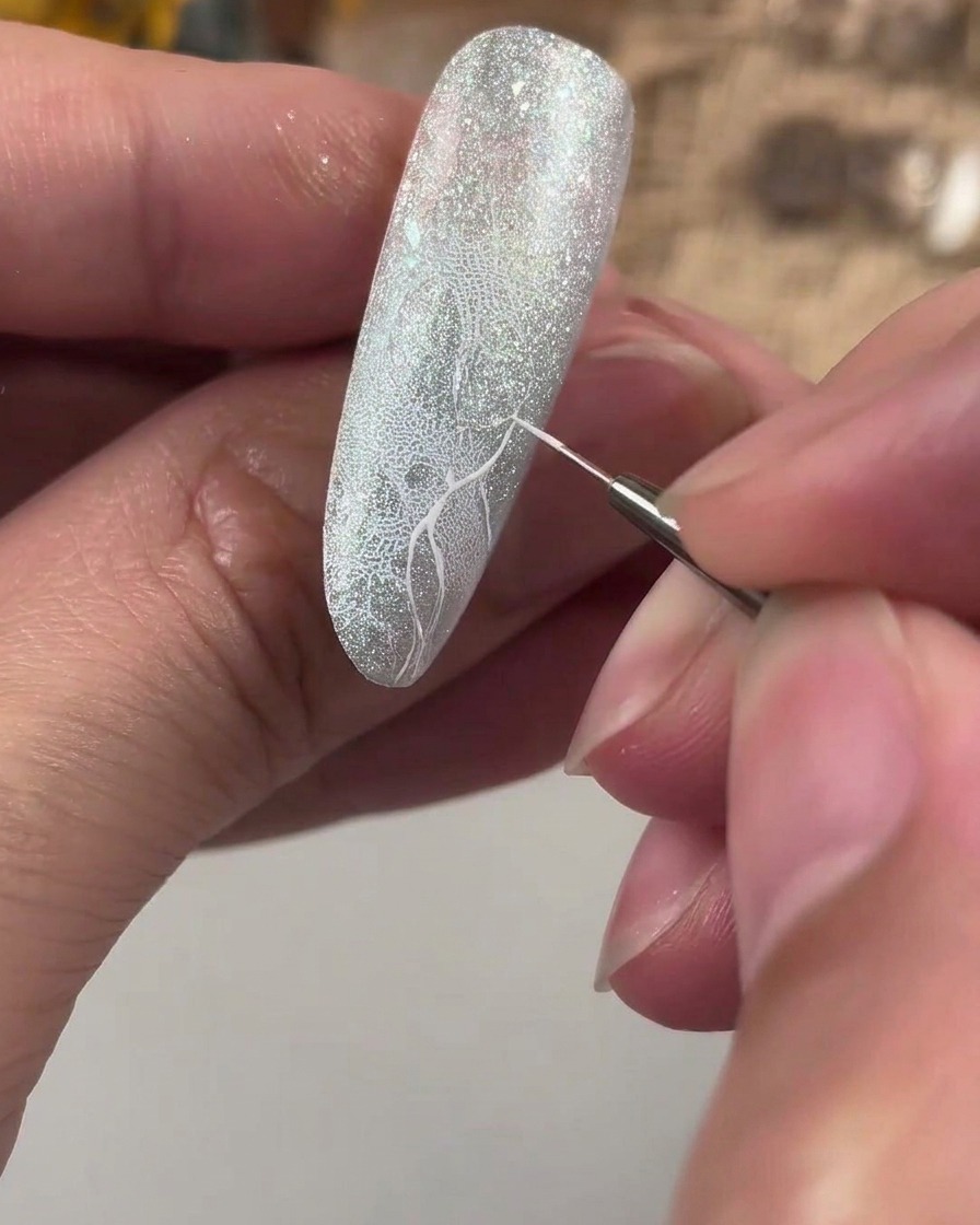 Un pinceau de détail fin dessinant une croix de repère blanche sur un ongle pailleté texturé.