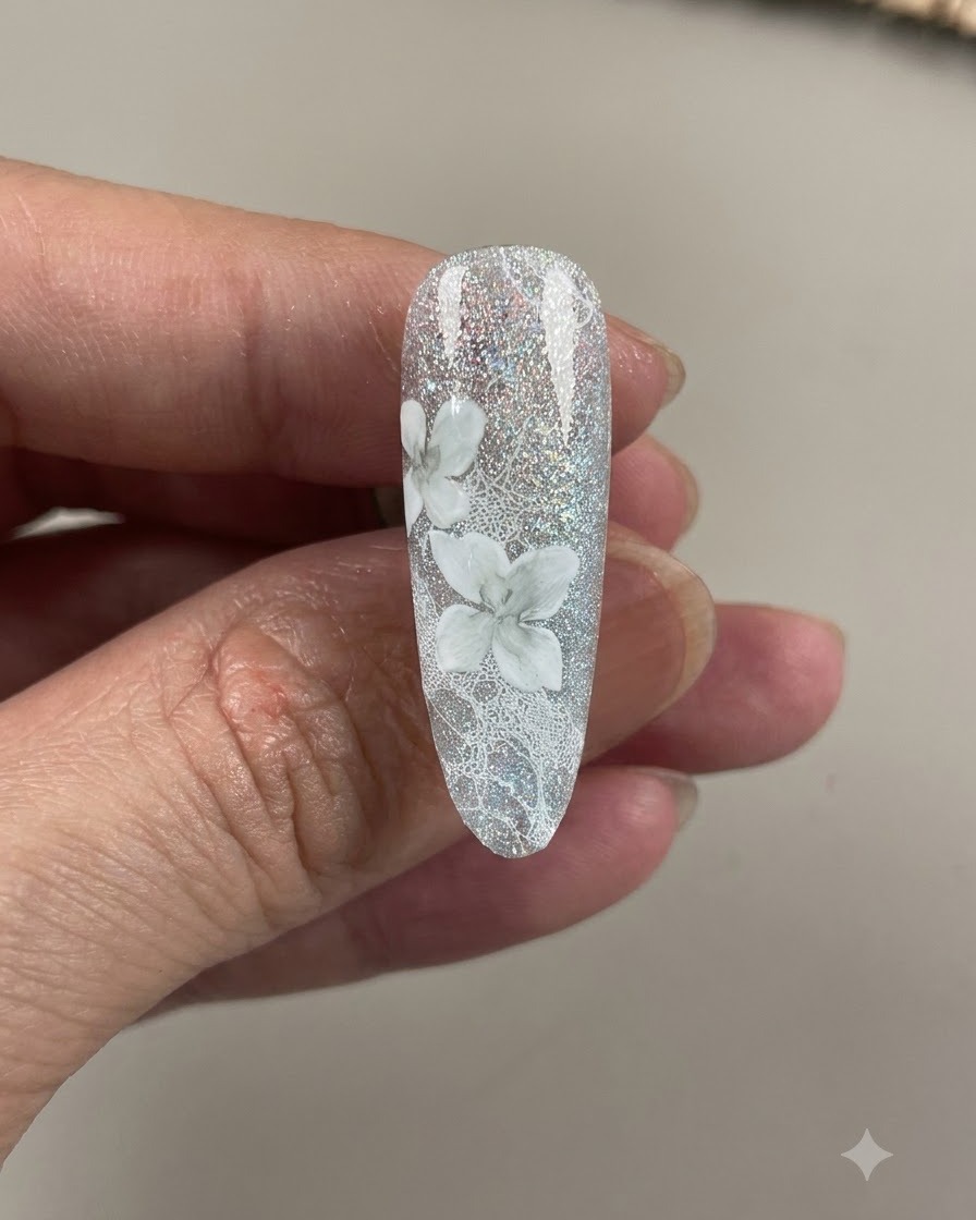 Un ongle terminé montrant des fleurs de camélia blanches détaillées et ombragées sur un fond de dentelle gris-bleu scintillant.