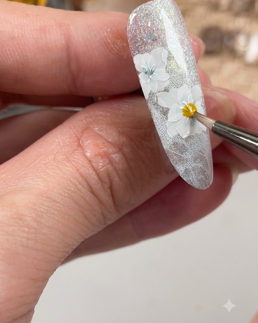 Un pinceau à ongles appliquant un top coat mat spécifiquement sur les détails de fleurs blanches façonnés sur longle.