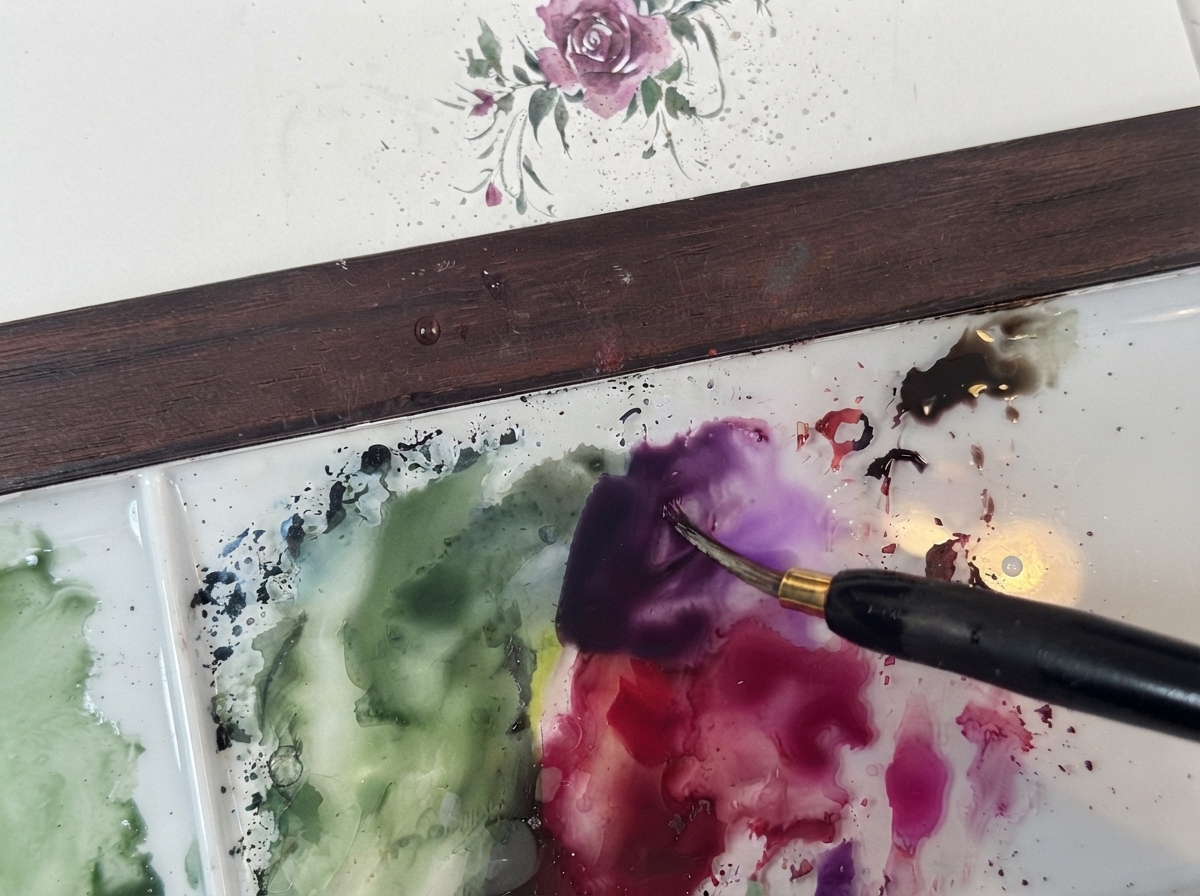 Un pinceau fin mélangeant un pigment violet foncé sur une palette blanche à côté de touches vertes.