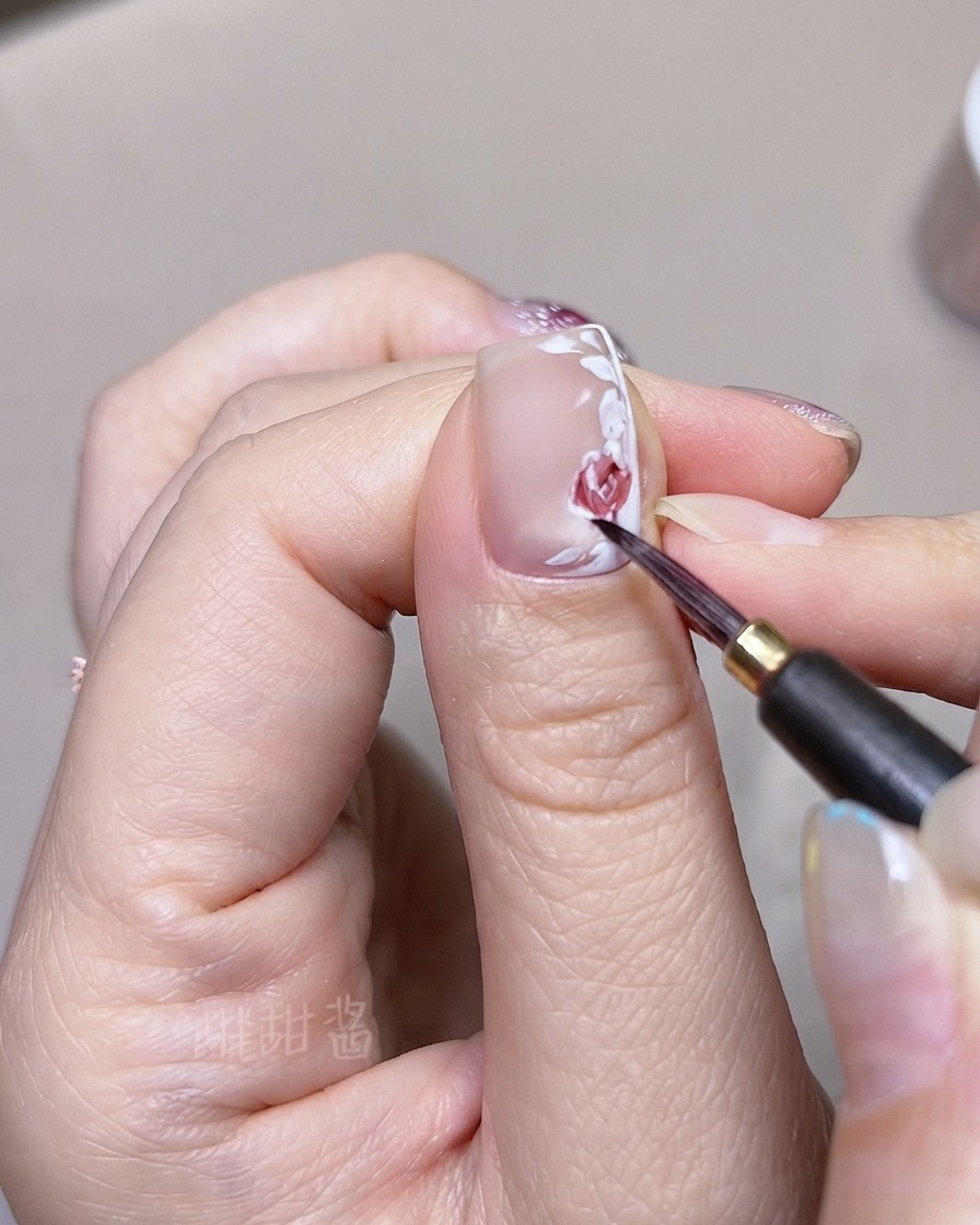 Un pinceau fin superposant des tons daquarelle rouge plus foncés sur des pétales de rose et des feuilles peints individuellement sur un ongle.
