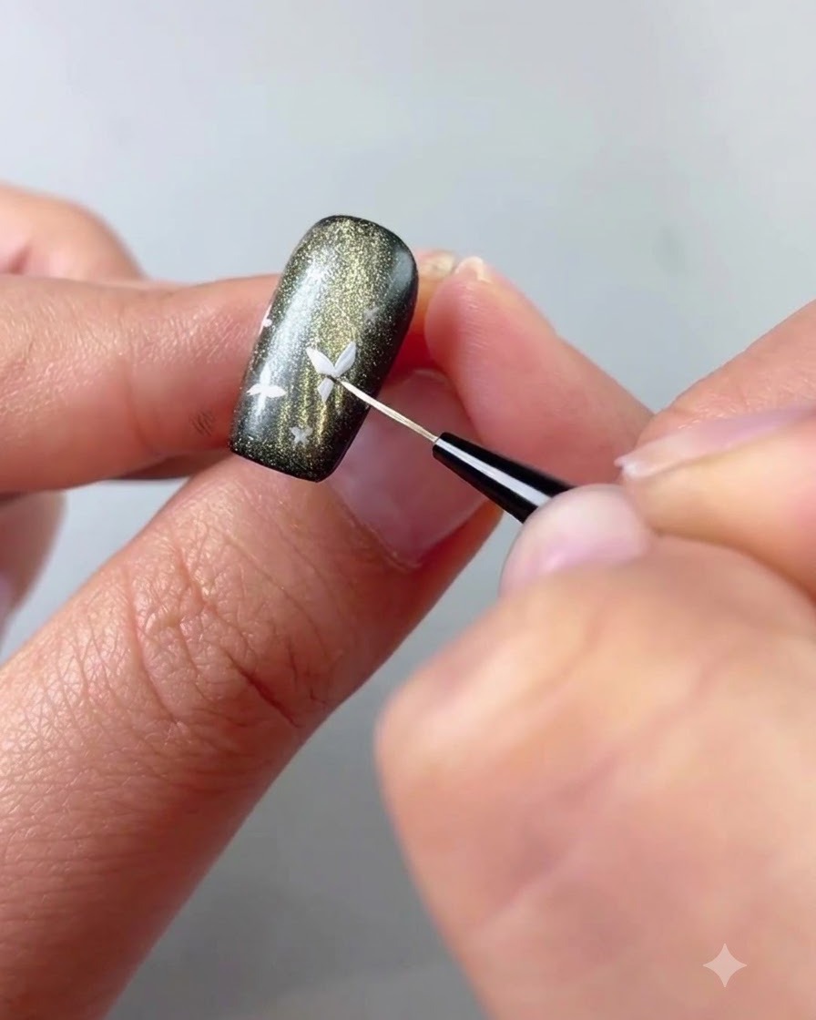 Un gros plan dun pinceau fin de détail appliquant du gel blanc pour peindre un petit détail de papillon sur un ongle cat-eye sombre.