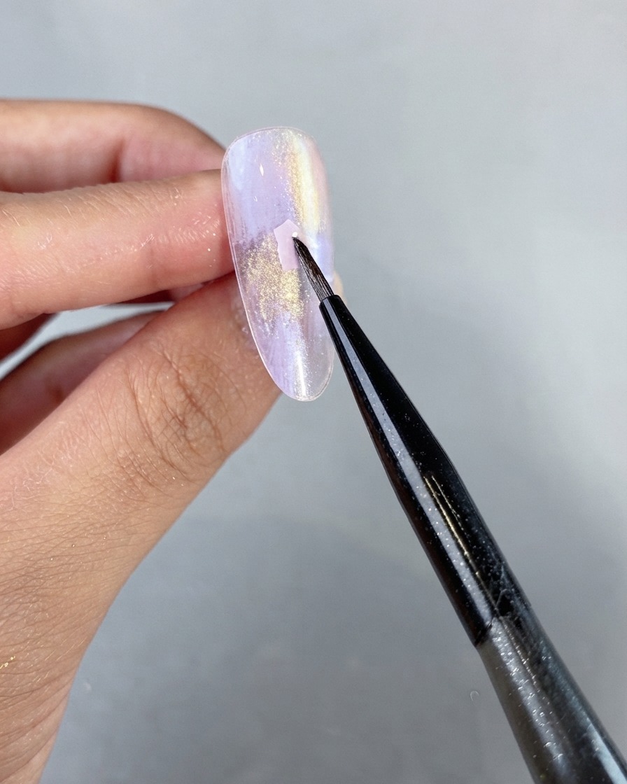 Un pinceau fin de nail art plaçant délicatement un morceau de coquillage rose sur la surface dun ongle en gel nacré violet non catalysé.