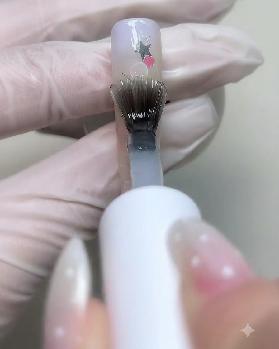 Un pinceau appliquant du gel de base transparent sur des ongles décorés de sequins détoiles et de bulles.