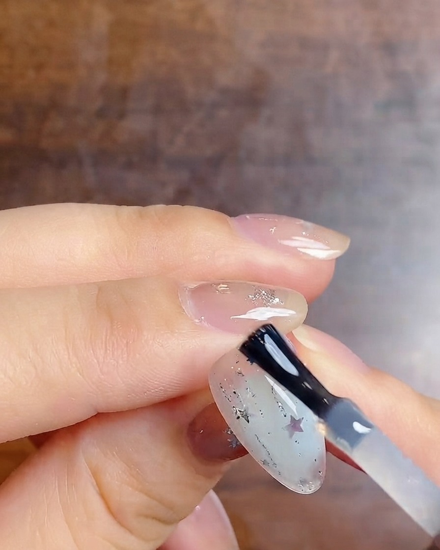 Un pinceau à ongle appliquant un top coat brillant sur un ongle dégradé décoré de paillettes et détoiles.