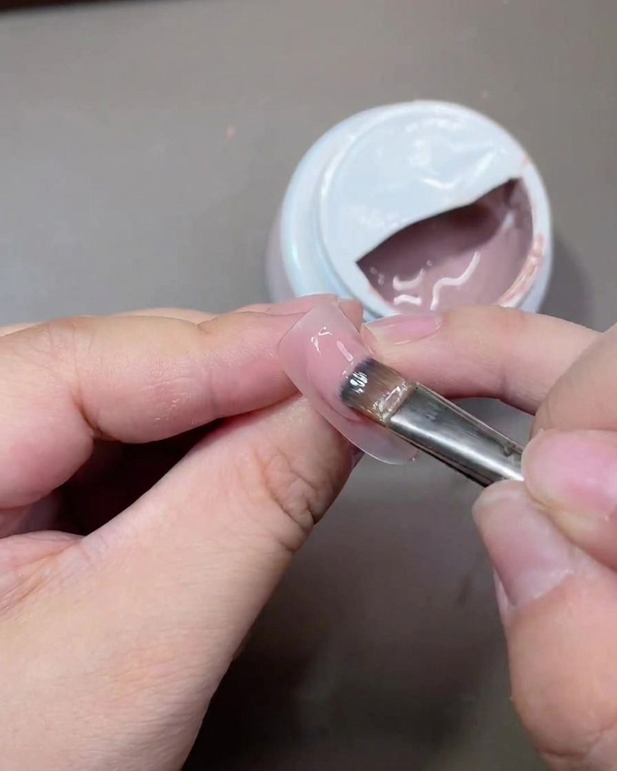 Un pinceau à gel appliquant une fine couche de vernis nude translucide sur la surface dun ongle.