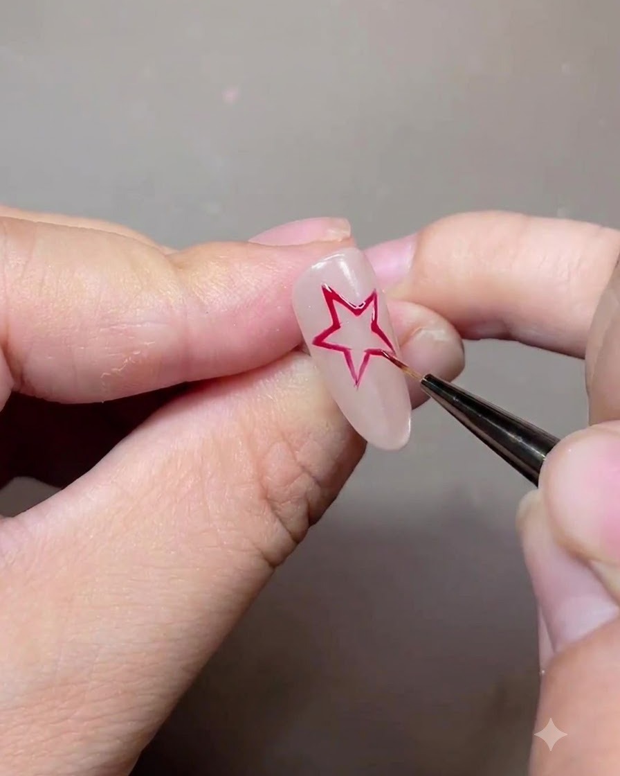 Un pinceau fin dessinant le contour dune étoile à cinq branches rouge sur un ongle en gel nude.