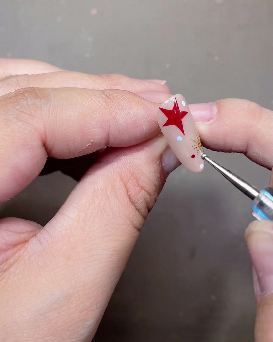 Un outil à pois plaçant de minuscules points blancs et rouges sur un ongle en gel nude décoré.