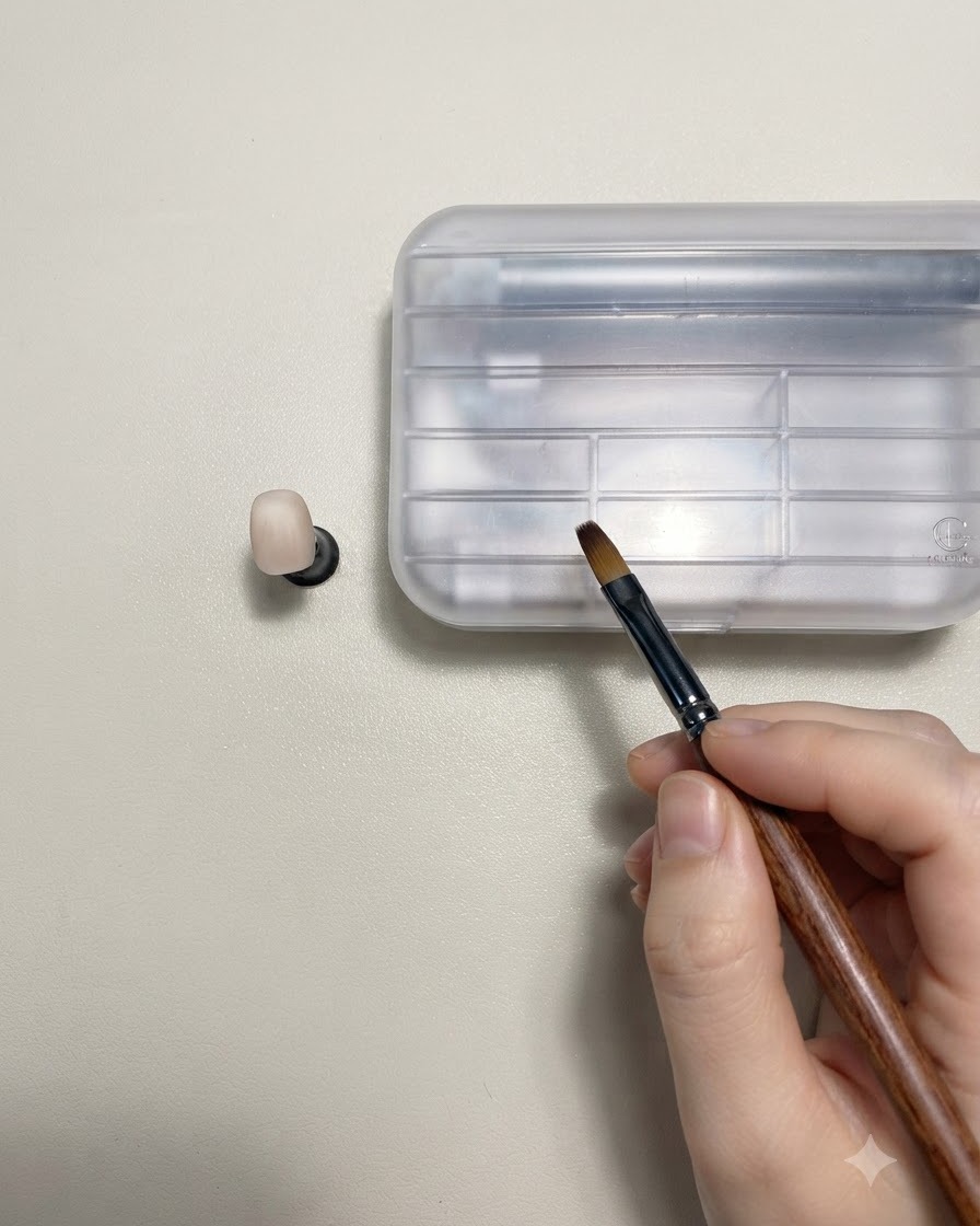 Une main tenant un pinceau de nail art au manche en bois à côté dun faux ongle de couleur nude sur un support.