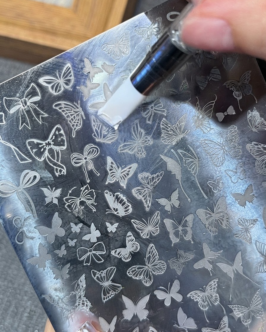 Un outil étalant du gel de stamping blanc sur un motif de papillon gravé sur une plaque métallique.