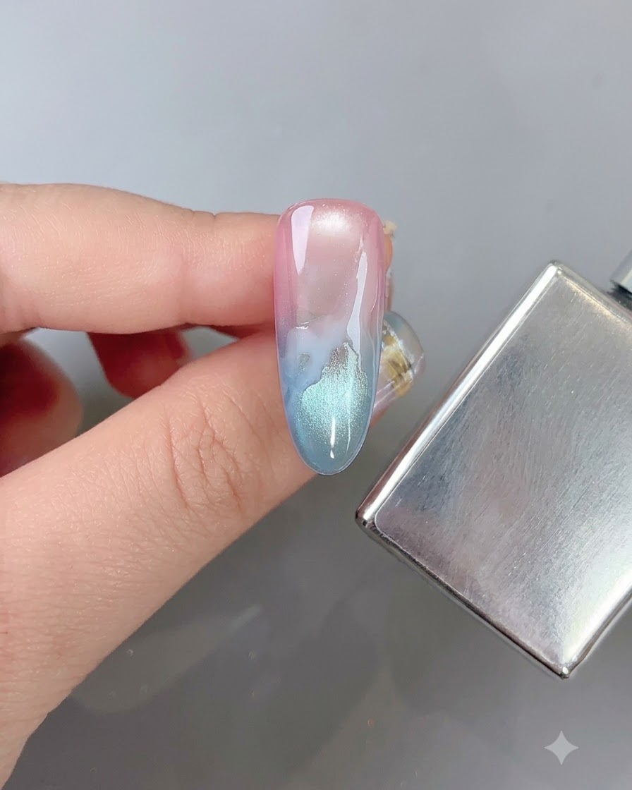 Un pinceau à ongle floutant doucement les bords dun motif de nuage blanc sur un ongle en dégradé multicolore à côté dun bâtonnet aimanté.