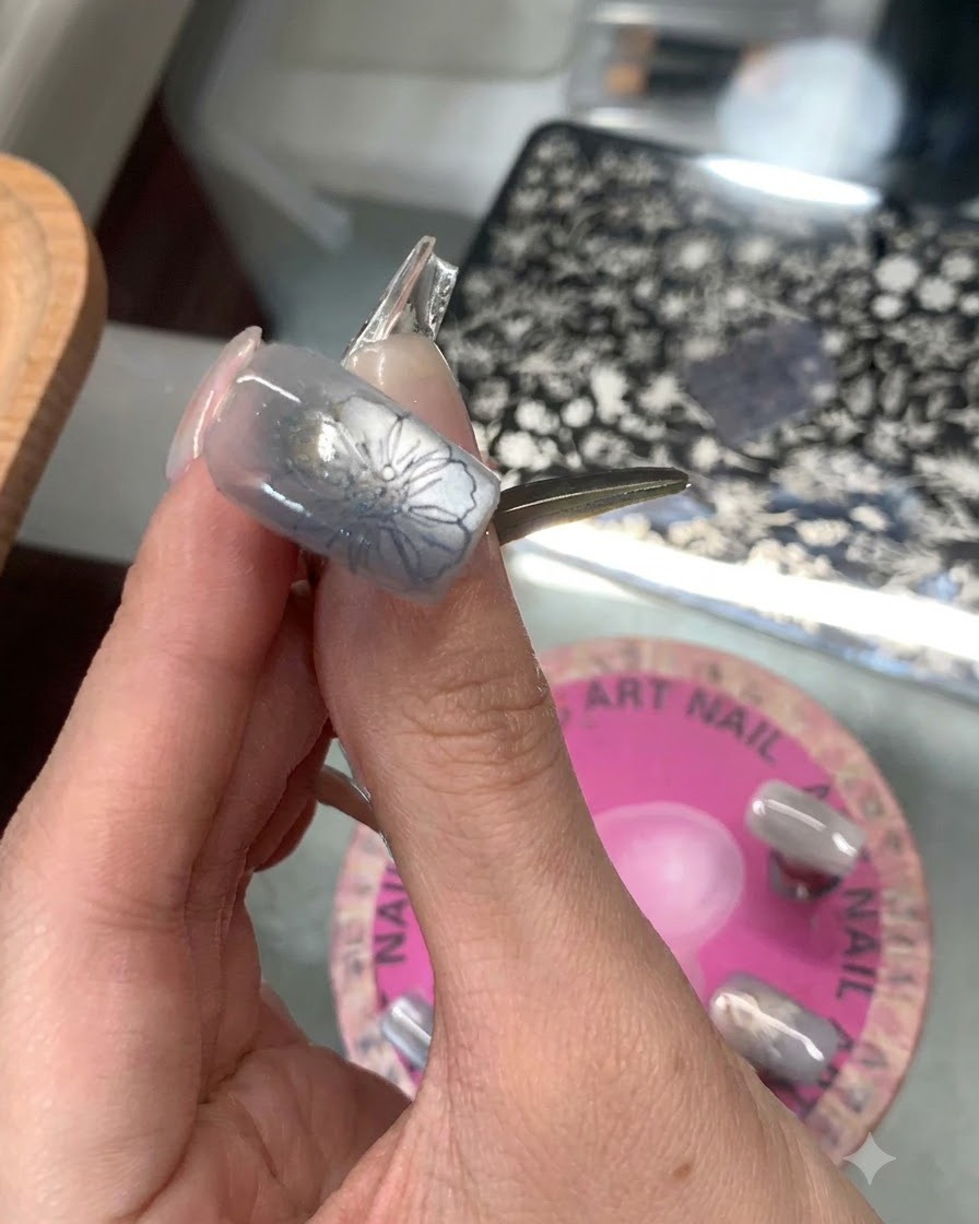 Un tampon en silicone transparent pressant un motif floral bleu sur le bout dun pouce manucuré bleu grisâtre.