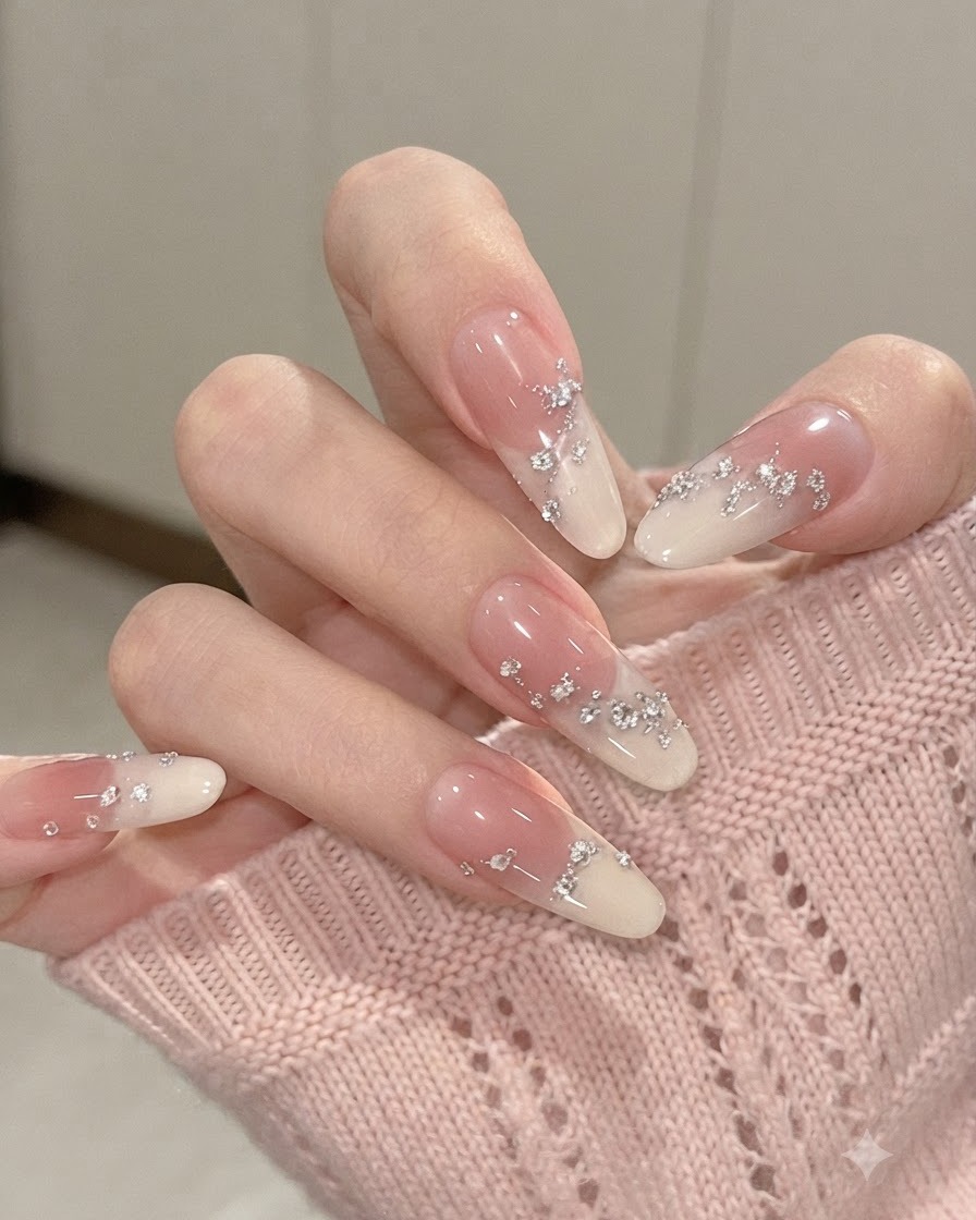 Gros plan sur des ongles en amande dégradés aux doigts légèrement pliés, mettant en valeur les feuilles dargent et les petits strass.