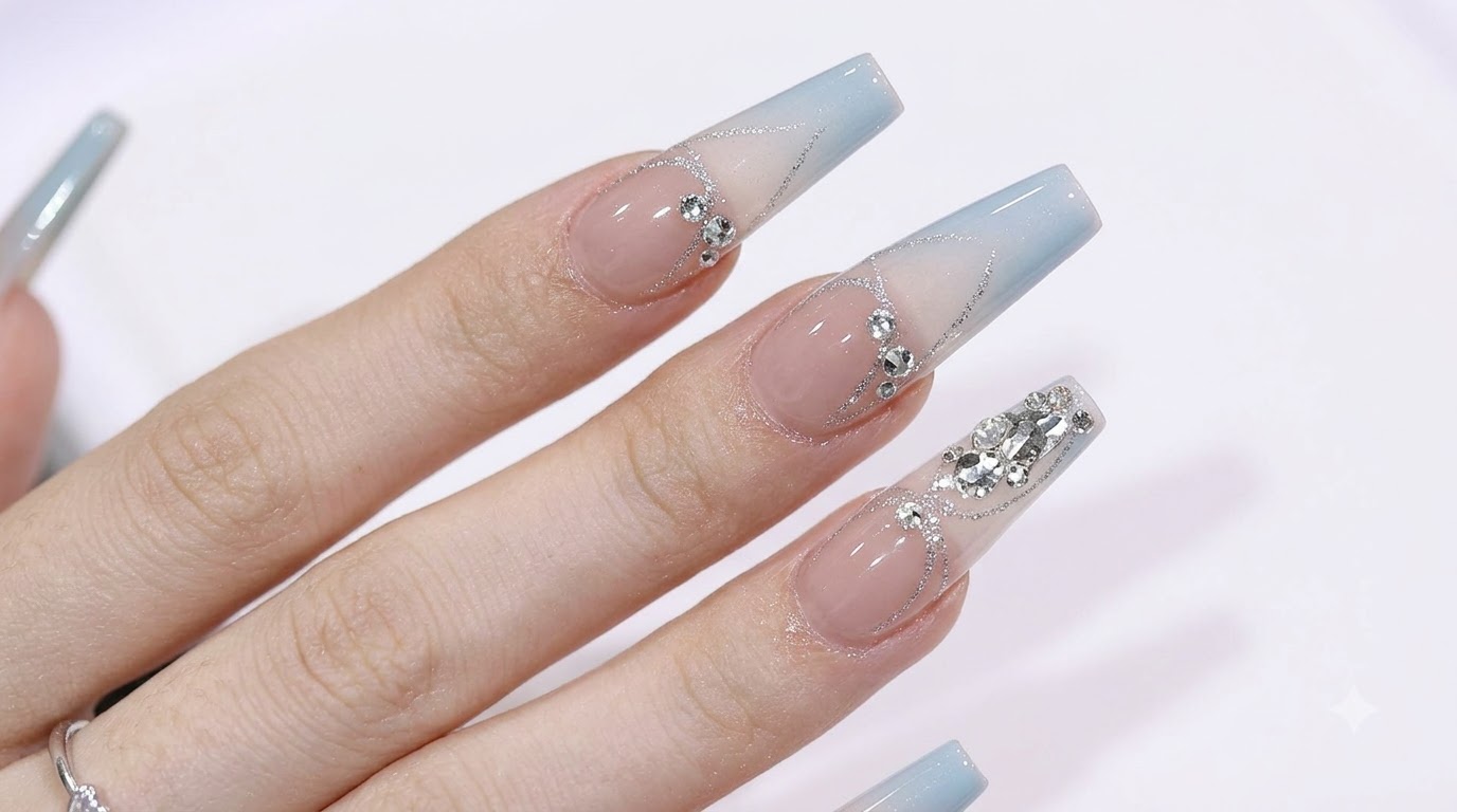 Ongles longs en forme de coffin arborant un dégradé doux du bleu au nude, rehaussés de strass étincelants et de détails en paillettes argentées.