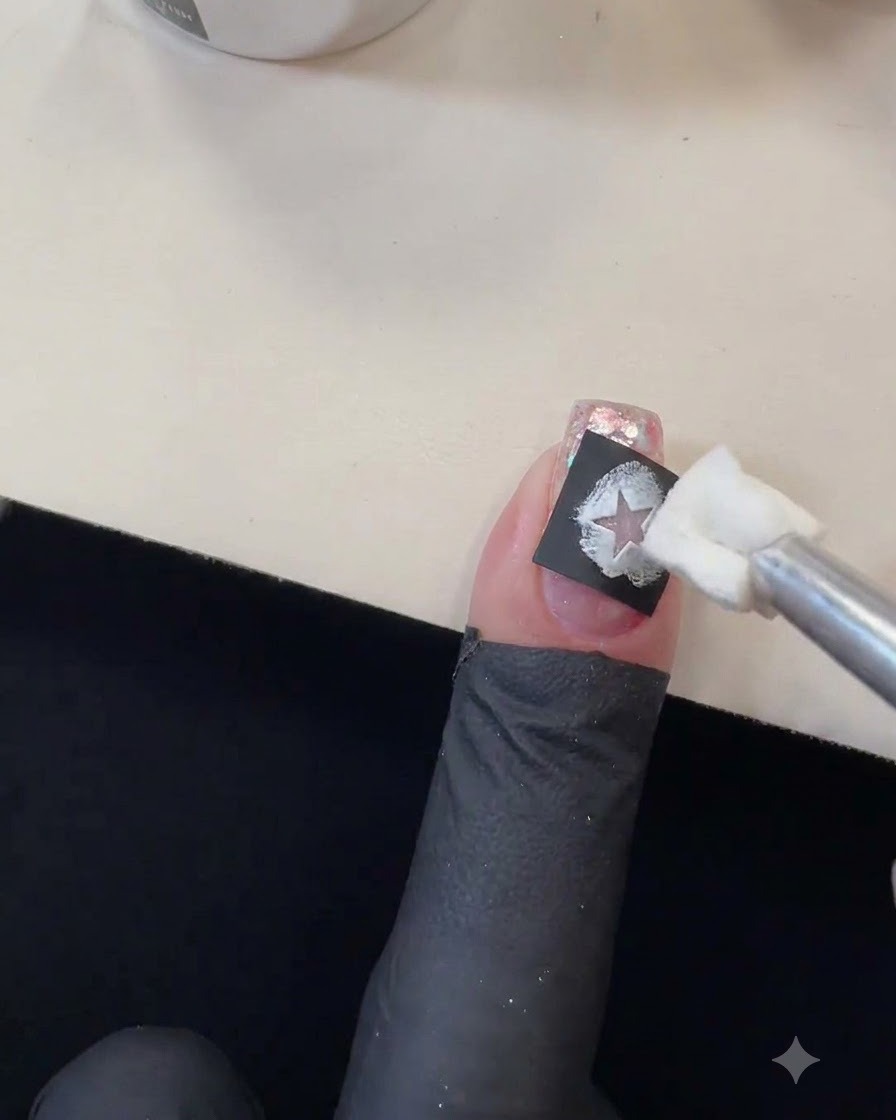Applicateur éponge tamponnant du gel polish blanc dans la découpe dun pochoir détoile noir sur un ongle pailleté.