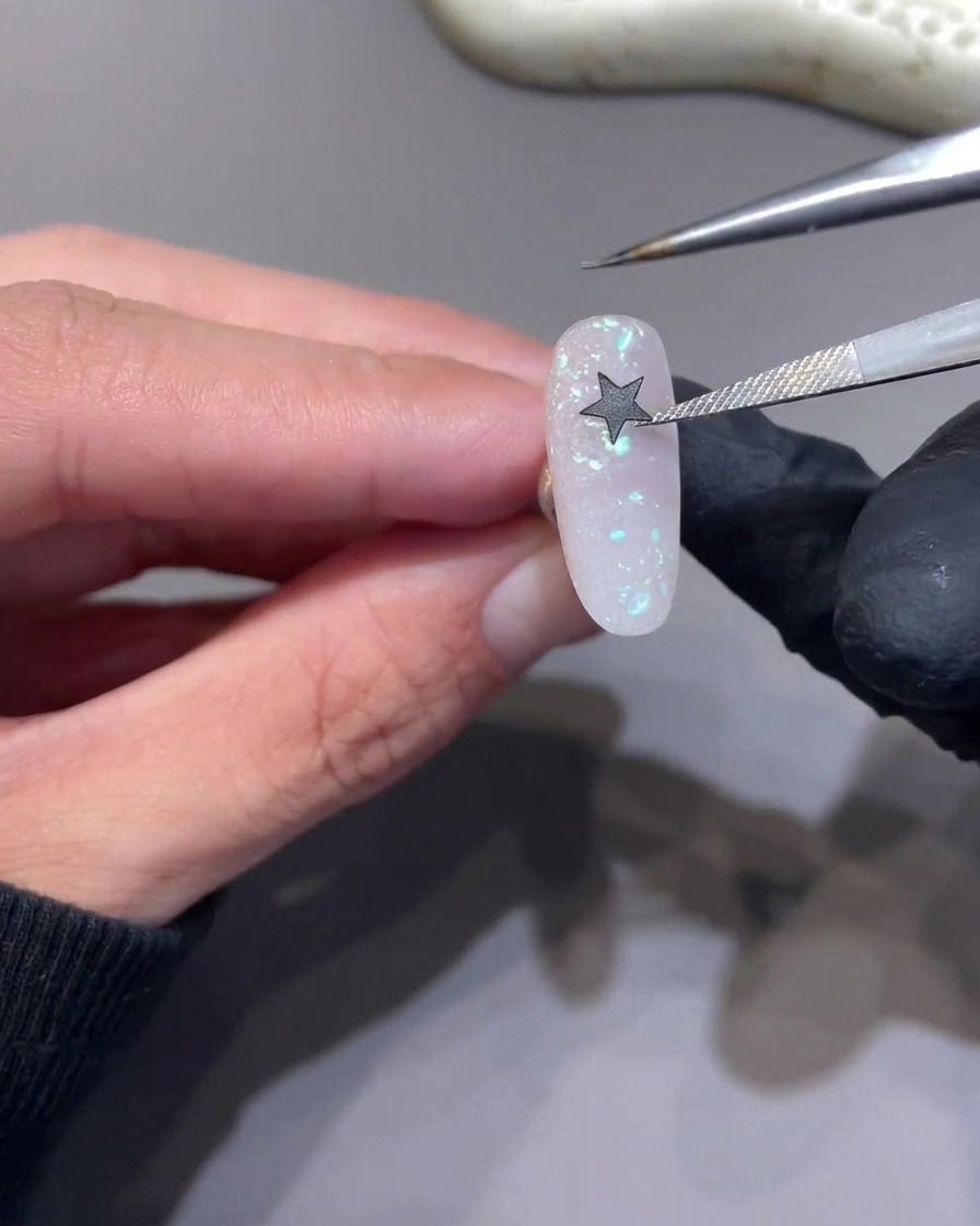 Une pince à épiler positionnant un autocollant étoile noire sur la surface dun ongle rose pailleté.
