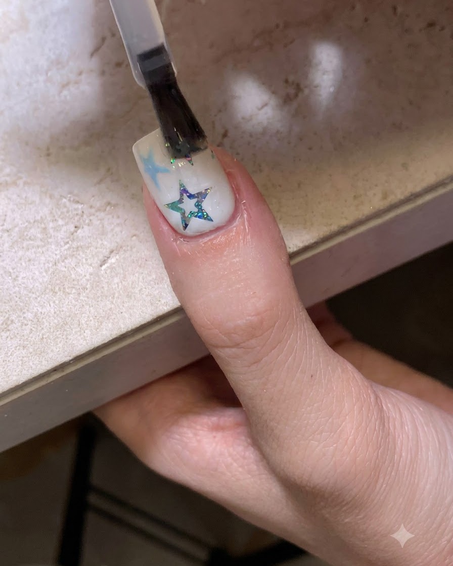 Un pinceau appliquant une couche de finition transparente et brillante sur un ongle décoré détoiles et de paillettes.