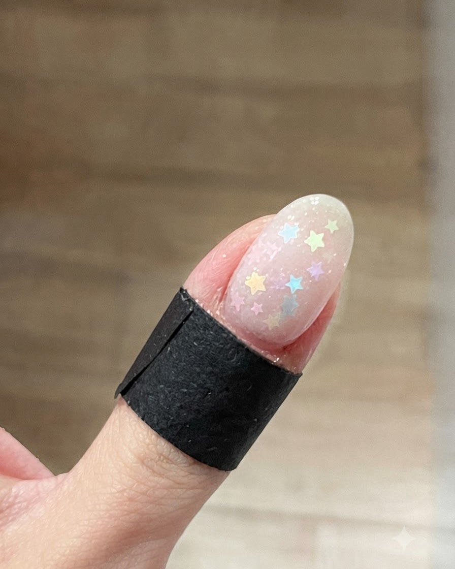 Gros plan sur un ongle ovale parsemé de paillettes étoiles multicolores pastel sur une base en gel humide transparente.