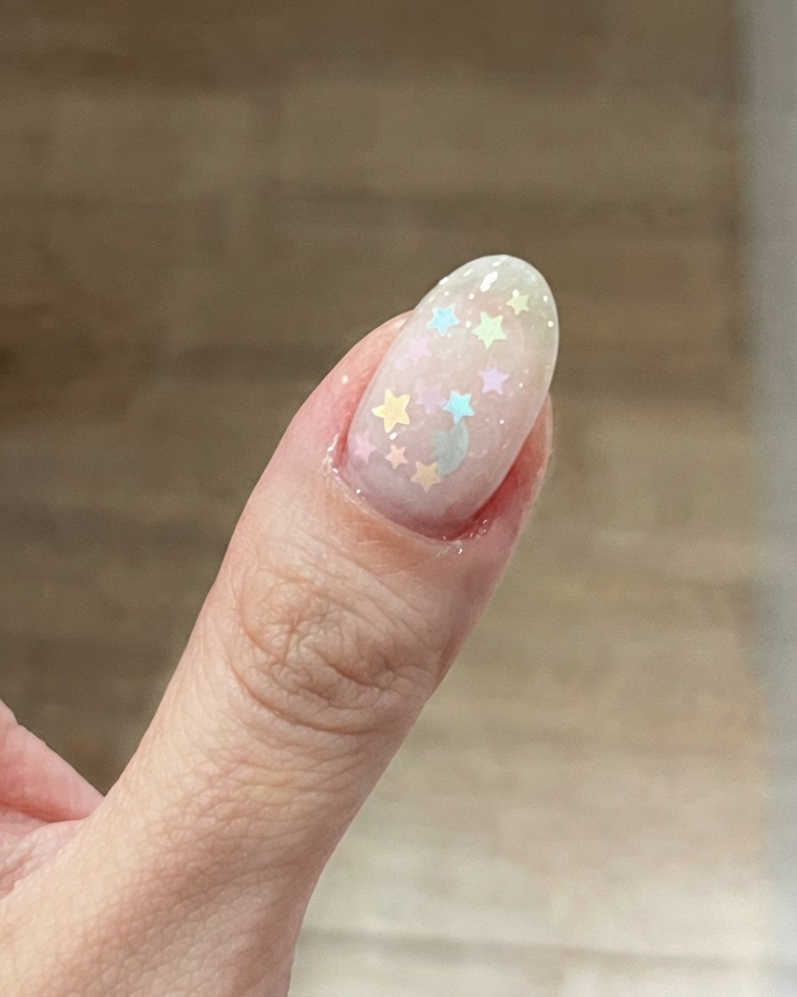 Gros plan sur un ongle de pouce doté dune base rose laiteux sur laquelle sont disposées des paillettes étoiles pastel colorées.