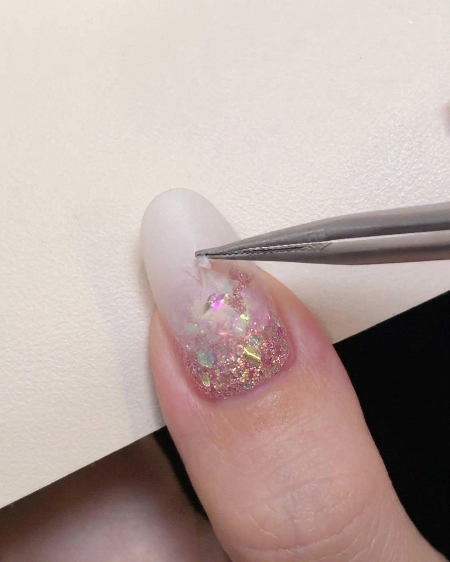 Une pince de précision retirant délicatement un autocollant détoile dun ongle à dégradé blanc laiteux pour révéler les paillettes roses.