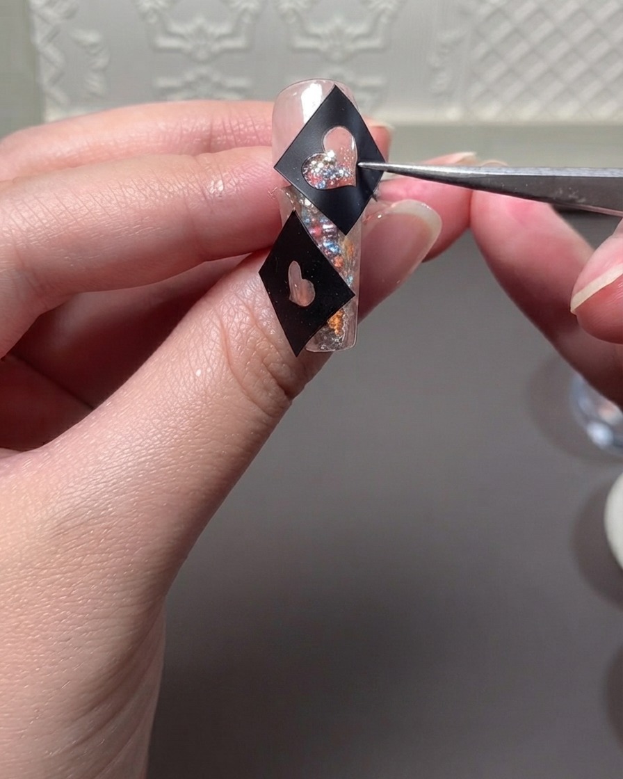 Pince tenant un pochoir cœur noir sur un ongle pailleté pendant lapplication du gel.