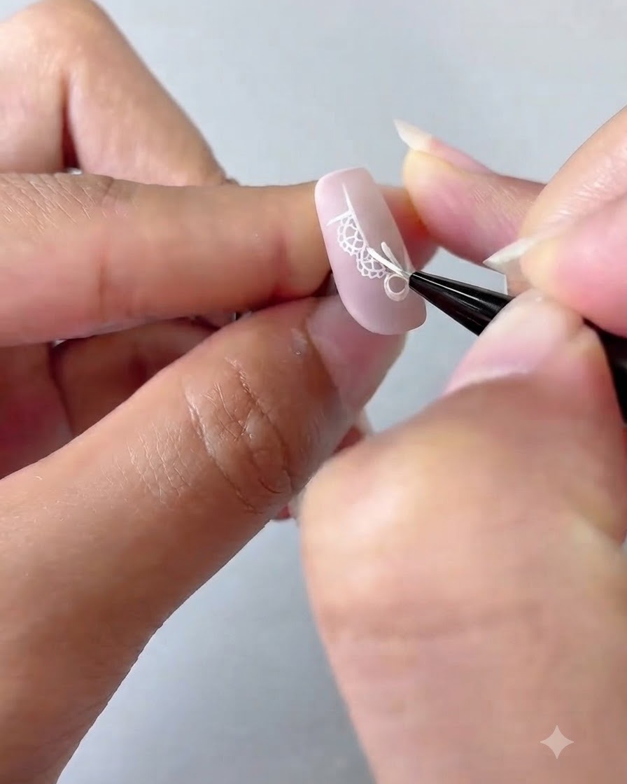 Un pinceau de nail art ajoutant des points blancs détaillés et une étoile à quatre branches sur un ongle dégradé rose.