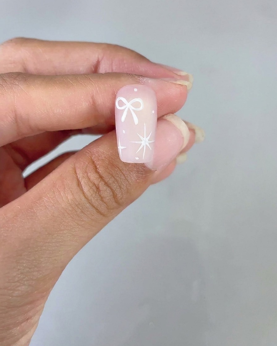 Un ongle décoré dun nœud blanc et dune étoile à quatre branches sur une base dégradée rose présenté pour visualisation.