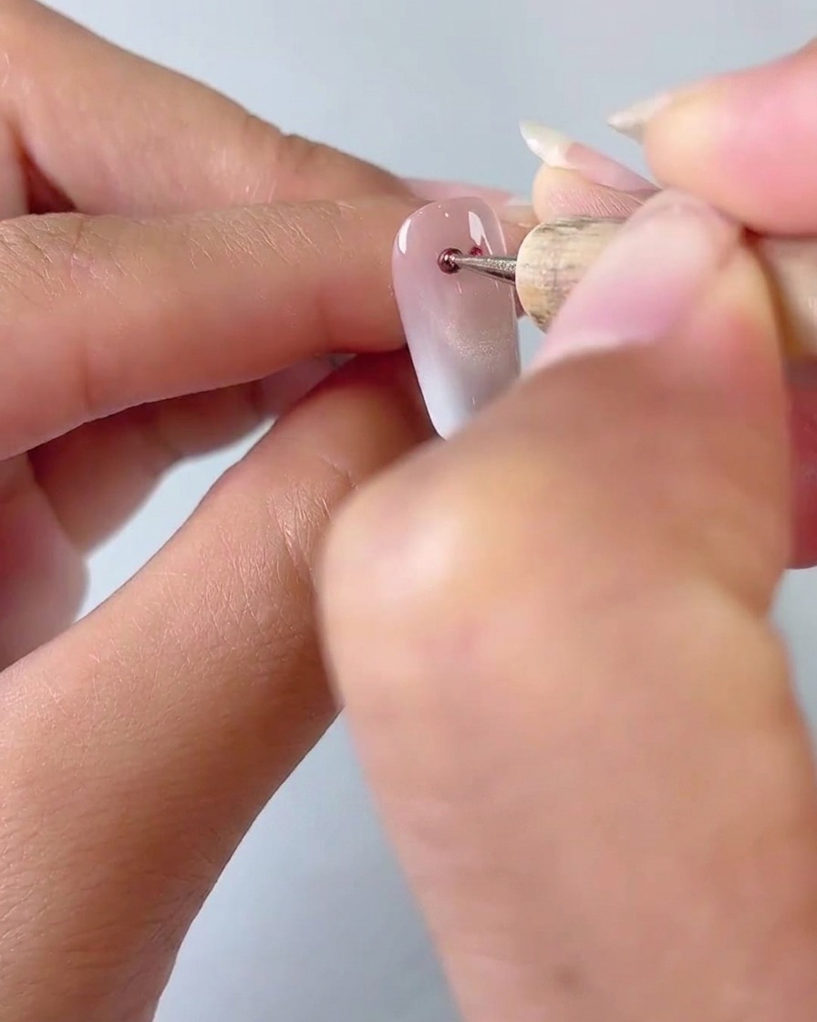 Gros plan dun outil de dotting appliquant deux petits points de gel cat eye rouge sur une base dongle dégradée laiteuse.