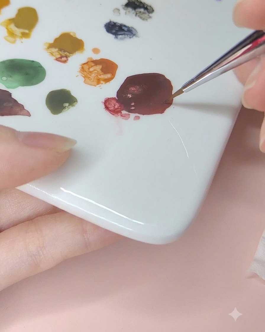 Un pinceau fin pour nail art ramassant de la peinture aquarelle rouge foncé sur une palette en céramique.