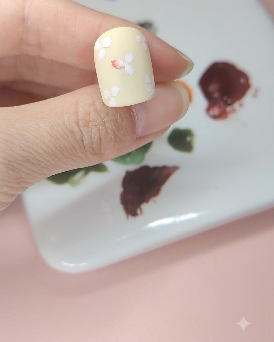 Un ongle jaune pâle avec des fleurs blanches où le centre de la fleur du milieu est teinté daquarelle rouge.