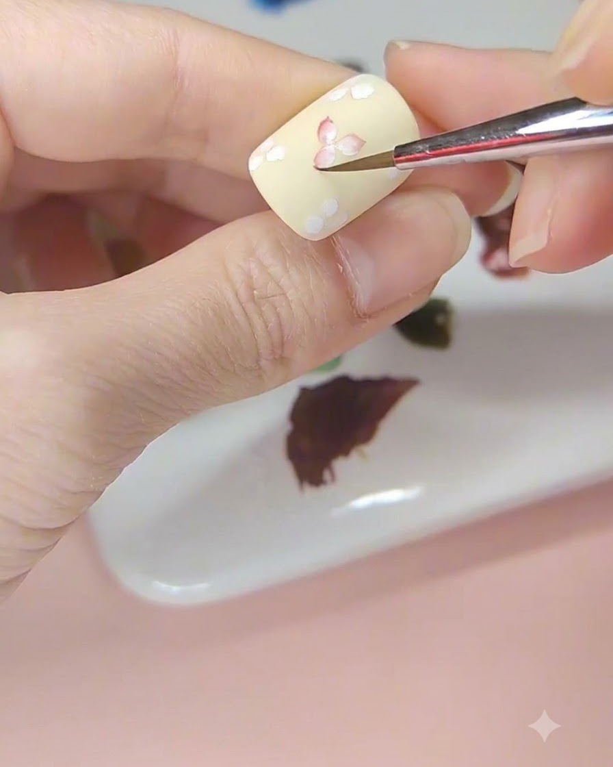 Un pinceau de nail art teintant le centre dune fleur blanche avec de laquarelle rouge sur un ongle jaune pâle.