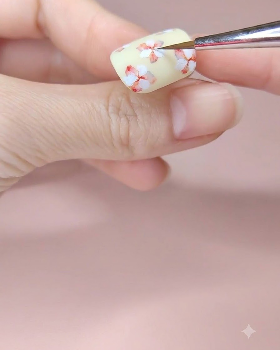 Un pinceau de nail art fin ajoutant une touche de pigment daquarelle rouge au centre dune fleur peinte en blanc sur un ongle jaune pâle.