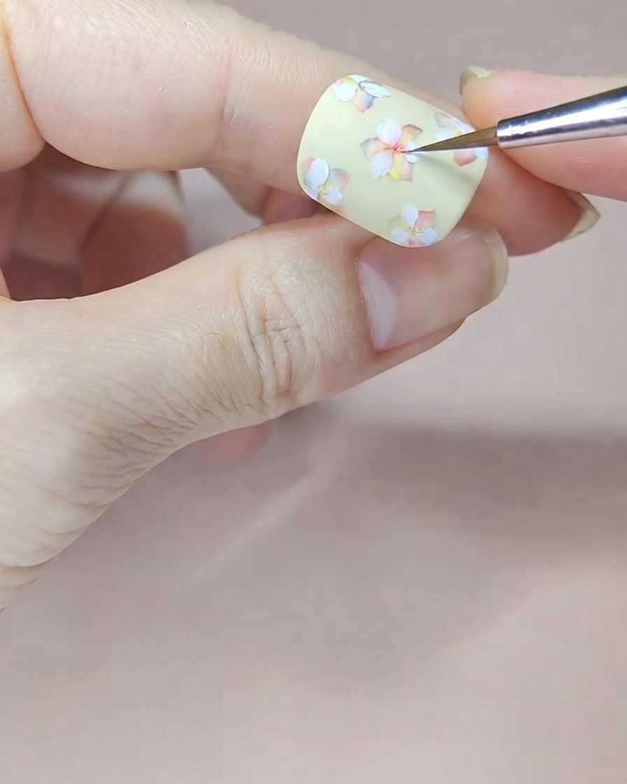 Un pinceau fin appliquant une teinte aquarelle rouge sur les centres des fleurs blanches sur un ongle jaune pâle.