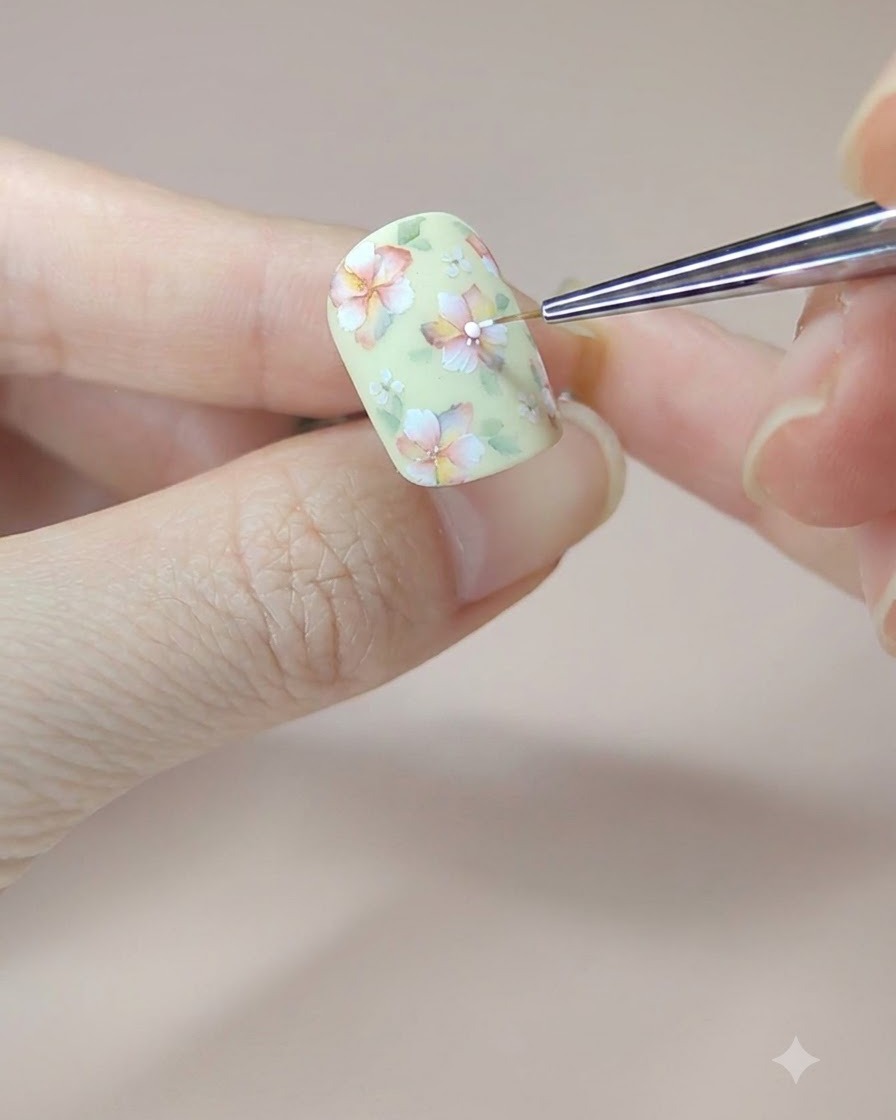 Un pinceau fin ajoutant les points blancs finaux dans les centres des fleurs sur un ongle jaune décoré.