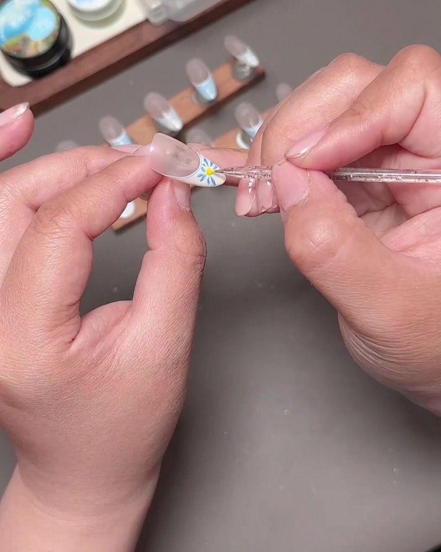 Un outil de dotting plaçant un point central jaune vif dans une fleur bleue peinte sur un ongle de manucure française.