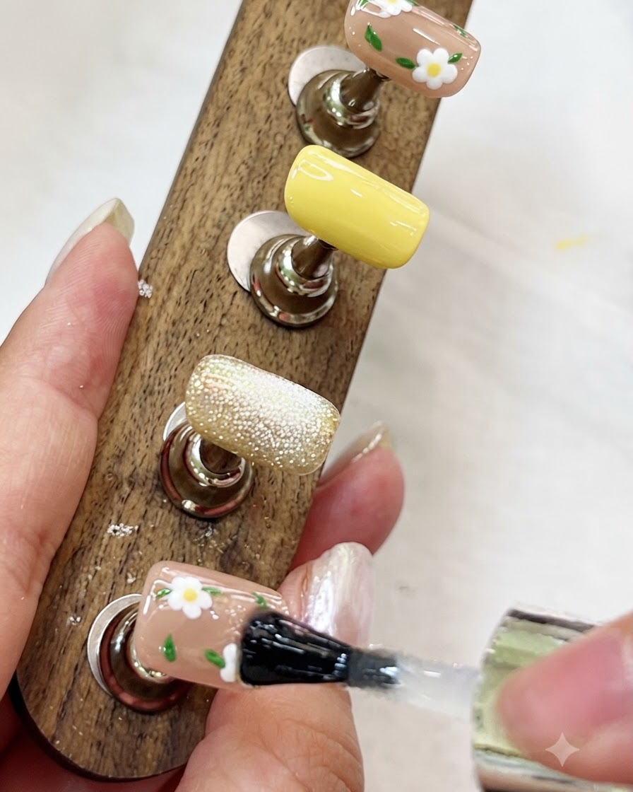 Un pinceau appliquant un top coat transparent et brillant sur un ongle peint de fleurs sur un présentoir en bois.