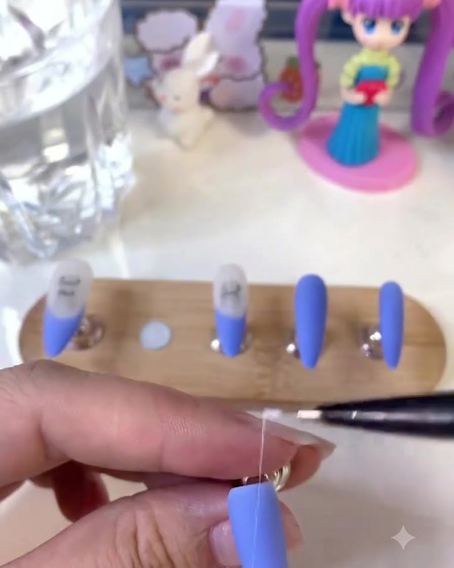 Une prothésiste ongulaire tenant un pinceau fin pour liner et dessinant des lignes blanches détaillées sur un ongle bleu mat.